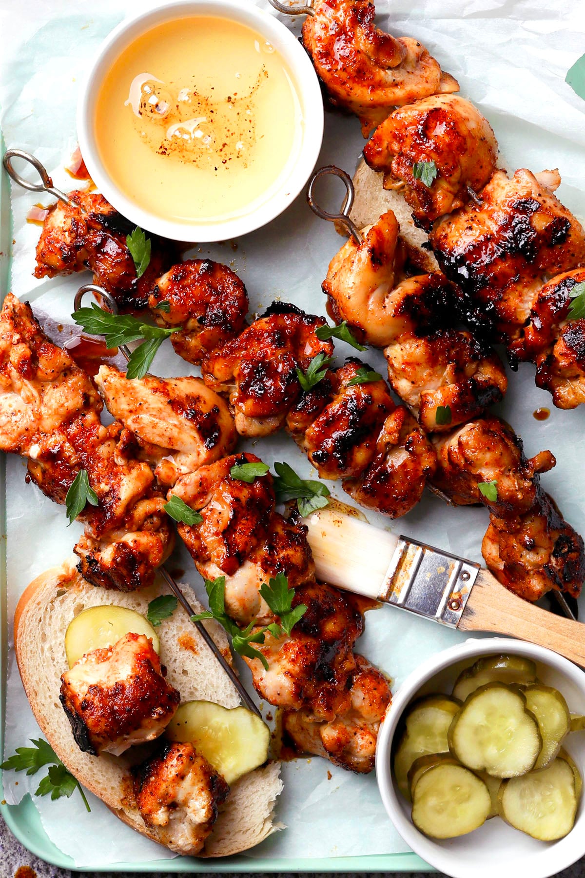 Grilled Nashville hot chicken skewers with a glazed finish are served on slices of bread with pickles, garnished with parsley. A brush, a bowl of honey, and a small bowl of pickles are also on the tray.