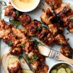Grilled chicken skewers on bread slices, garnished with parsley, next to a bowl of honey, a brush, and a small bowl of pickles on a parchment-lined tray.