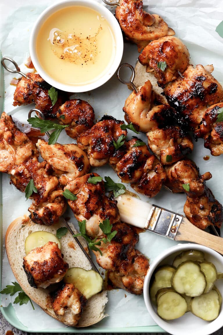 Grilled chicken skewers on bread slices, garnished with parsley, next to a bowl of honey, a brush, and a small bowl of pickles on a parchment-lined tray.