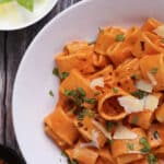A white bowl of rigatoni pasta in creamy tomato sauce, topped with fresh basil, red pepper flakes, and shaved parmesan cheese. A small bowl of basil leaves and parmesan is nearby on a rustic wooden surface.