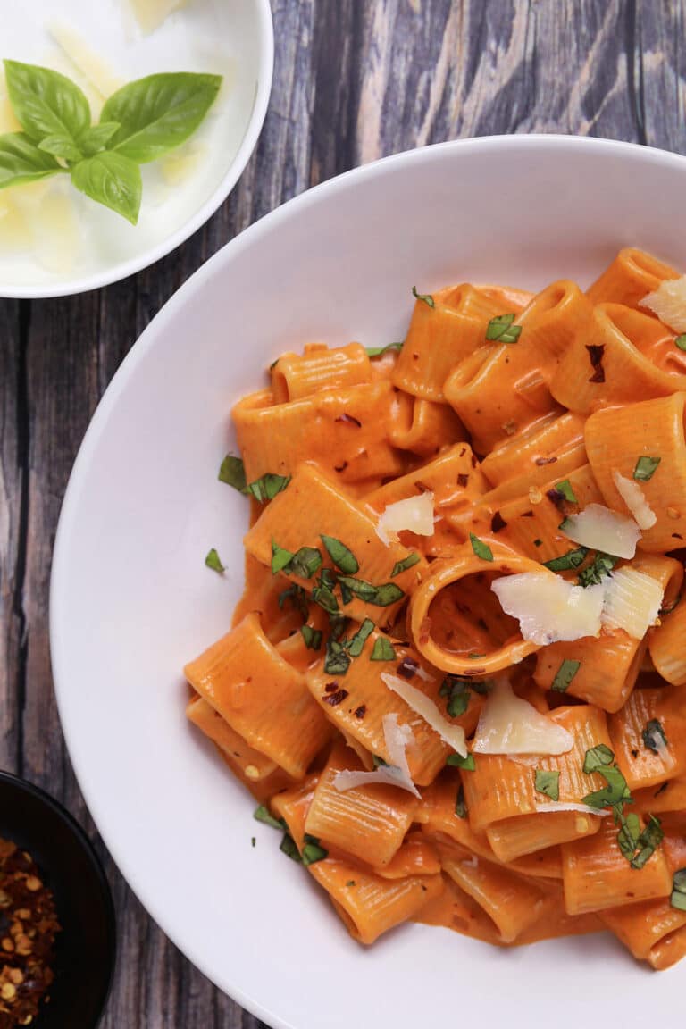 A white bowl of rigatoni pasta in creamy tomato sauce, topped with fresh basil, red pepper flakes, and shaved parmesan cheese. A small bowl of basil leaves and parmesan is nearby on a rustic wooden surface.