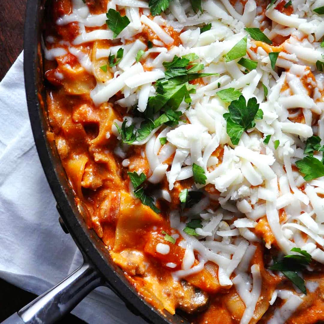 A close-up of a skillet filled with lasagna, topped with melted mozzarella cheese, tomato sauce, and fresh parsley, rests on a white cloth—a true farmgirl gourmet comfort dish.