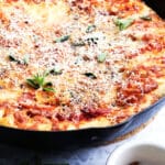 A freshly baked pizza in a black cast iron skillet, topped with tomato sauce, melted cheese, fresh herbs, and grated parmesan, with a small bowl of red pepper flakes on the side.