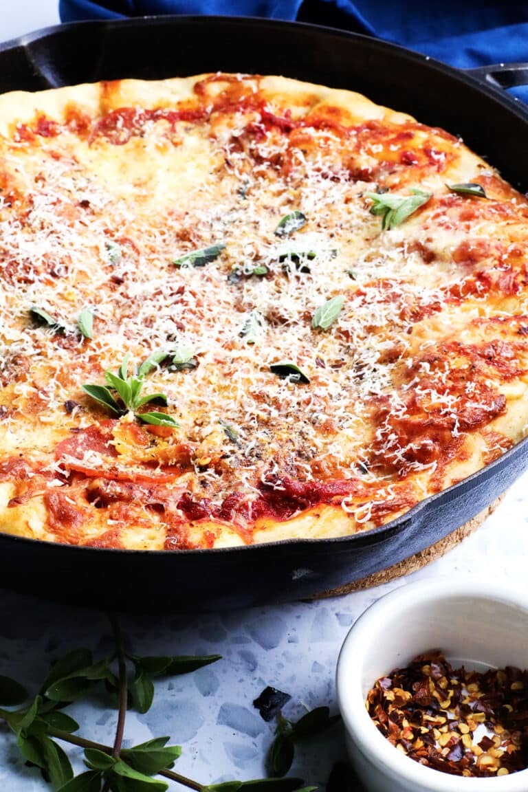A freshly baked pizza in a black cast iron skillet, topped with tomato sauce, melted cheese, fresh herbs, and grated parmesan, with a small bowl of red pepper flakes on the side.