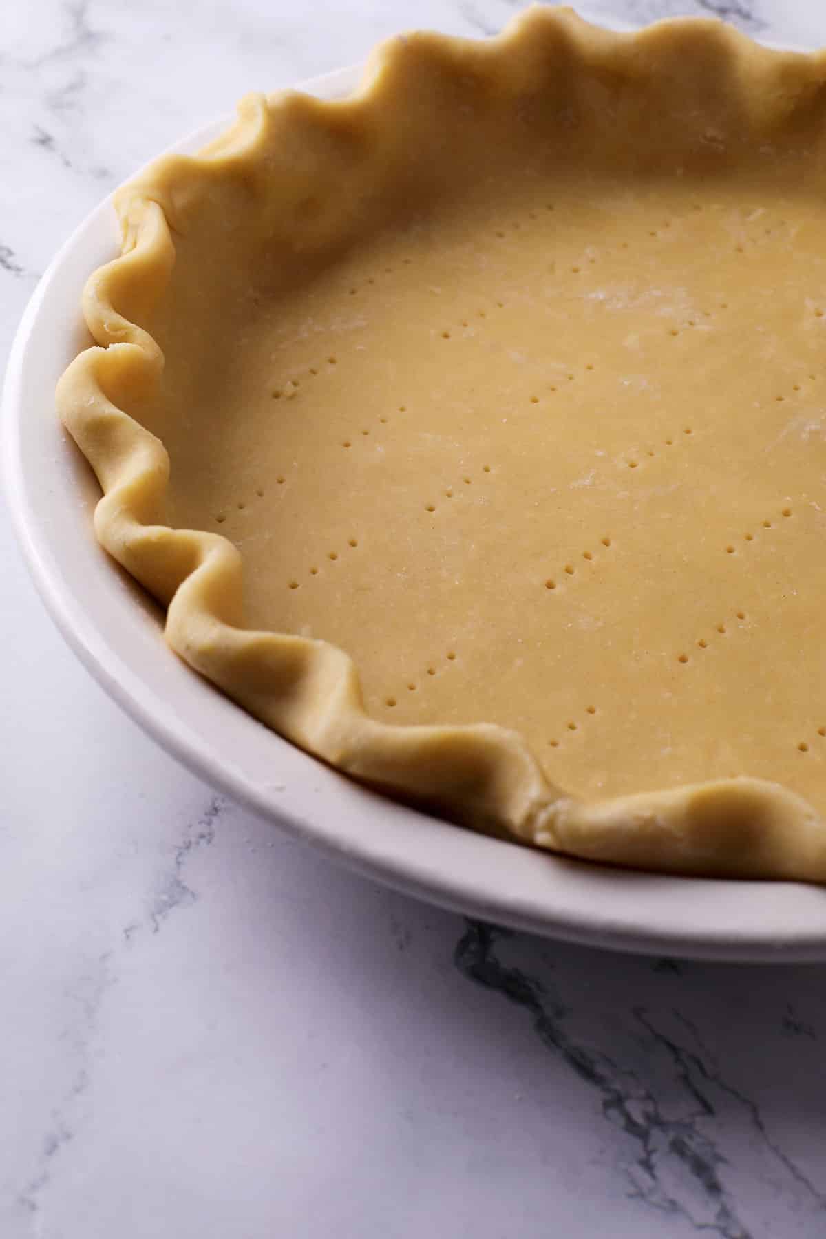 A close-up of an unbaked pie crust with crimped edges in a white pie dish, resting on a marble surface. Small holes are poked across the bottom of the dough.