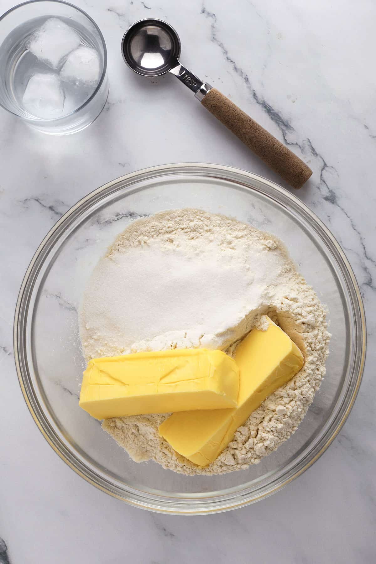 A glass bowl contains flour, sugar, and two sticks of butter on a marble surface. Nearby are a glass of ice water and a metal ice cream scoop with a brown handle.