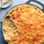 A casserole dish filled with baked macaroni and cheese, with a golden, crispy top and a serving scooped out. A spoon with cheesy residue rests on a nearby plate.