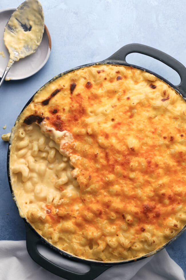 A casserole dish filled with baked macaroni and cheese, with a golden, crispy top and a serving scooped out. A spoon with cheesy residue rests on a nearby plate.