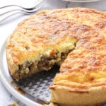 A golden-brown quiche with a slice missing, revealing a filling with mushrooms and cheese, sits on a plate. Stacked white plates and forks appear in the background.