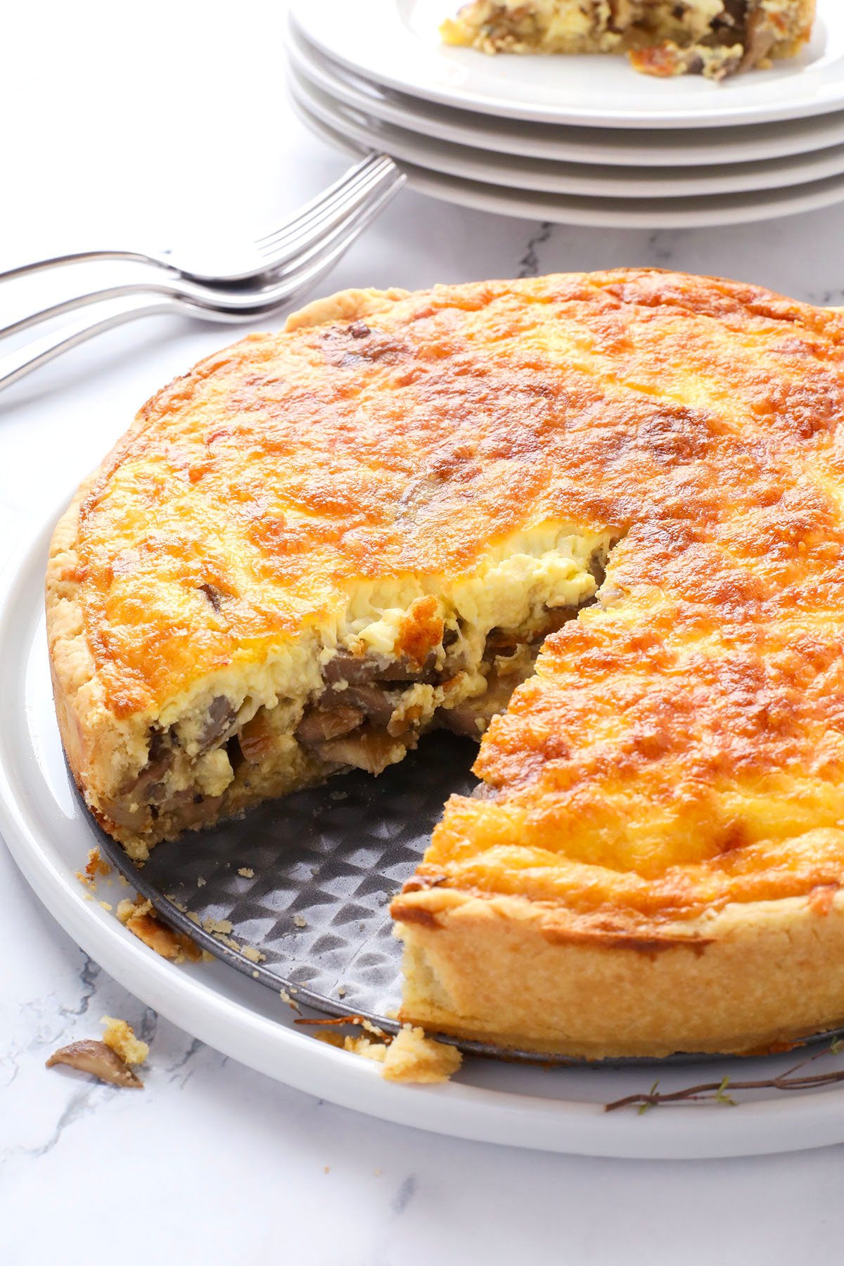 A golden-brown quiche with a slice missing, revealing a filling with mushrooms and cheese, sits on a plate. Stacked white plates and forks appear in the background.