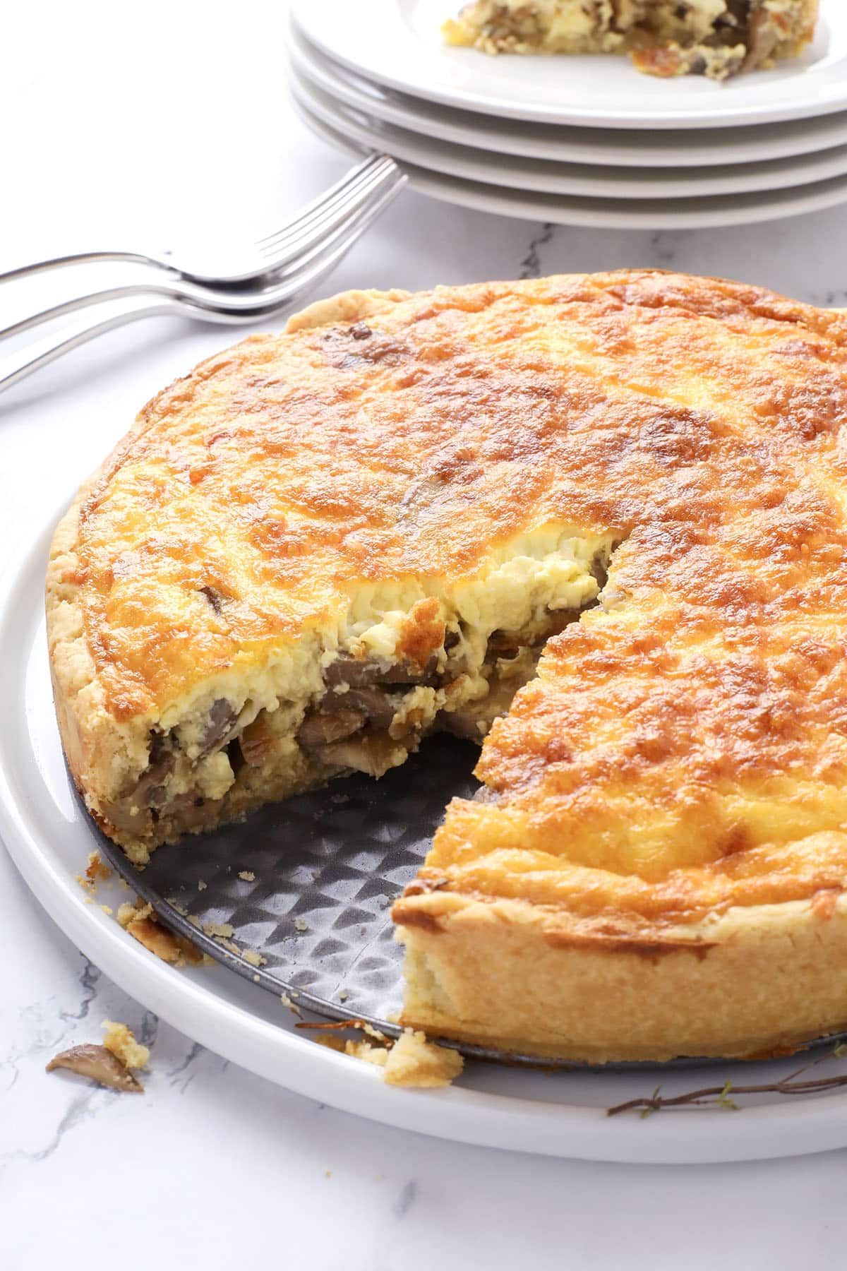 A golden-brown quiche with a slice removed, revealing a filling of mushrooms and cheese, sits on a plate. Stacked plates and forks are in the background.