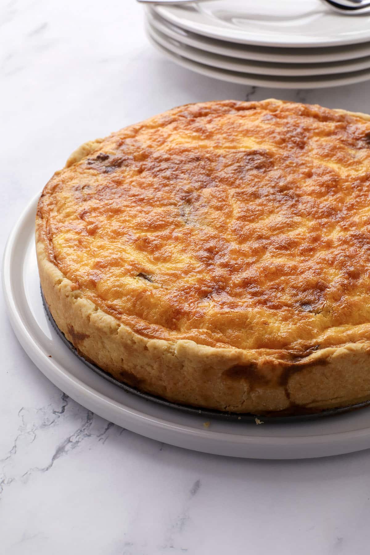 A golden-brown quiche with a flaky crust sits on a round white plate, with stacked white dishes and silverware in the background on a marble surface.