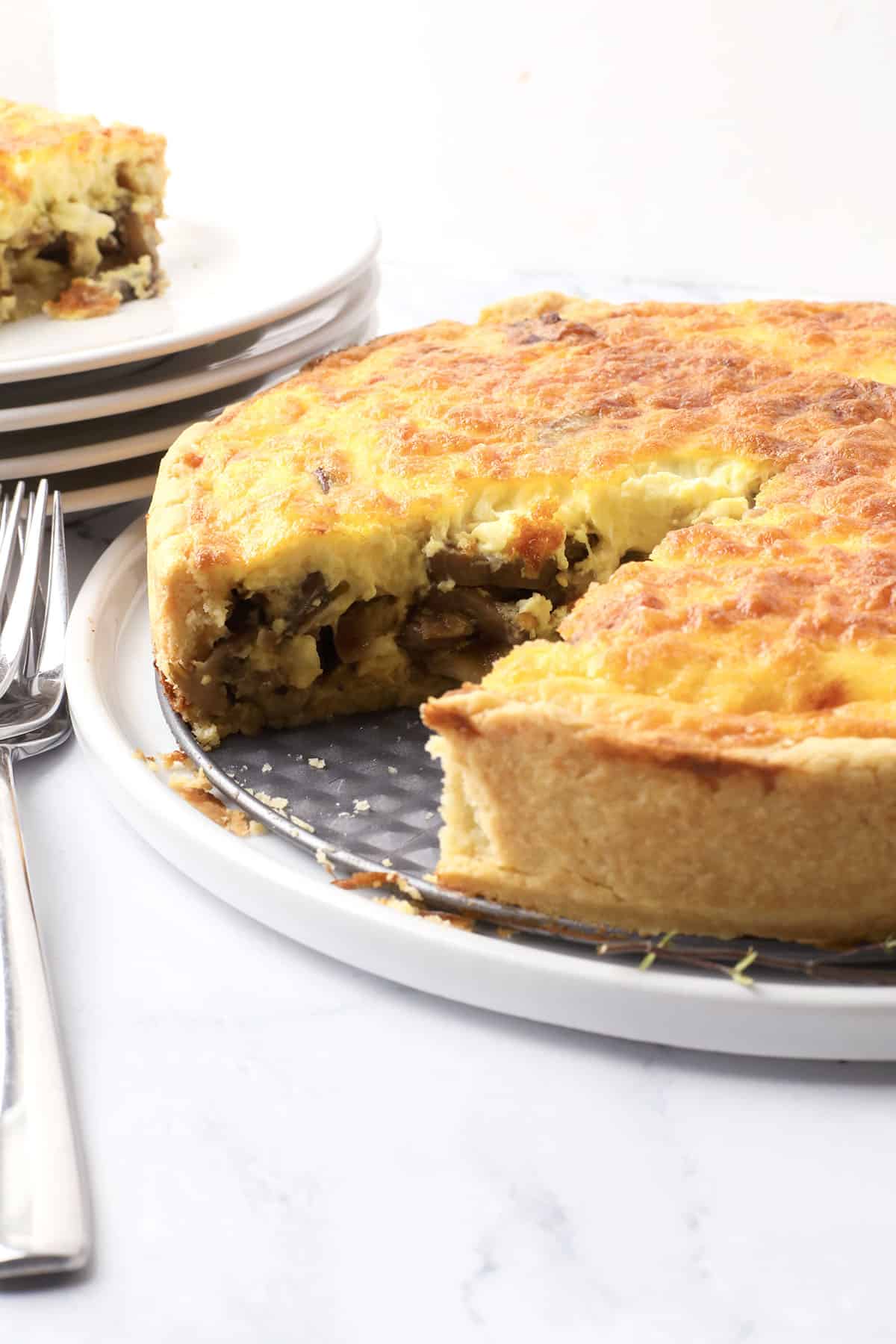 A golden-brown quiche with a slice removed, revealing a savory filling. The quiche sits on a round pan, next to stacked white plates and silverware on a marble surface.