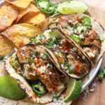 Three grilled fish tacos topped with chopped cilantro, jalapeños, cheese, and sauce, served with tortilla chips, lime wedges, and sliced jalapeños on a white plate with parchment paper, on a wooden board.