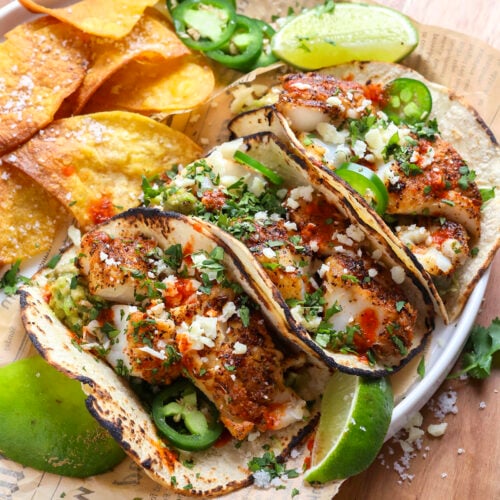 Three grilled fish tacos topped with chopped cilantro, jalapeños, cheese, and sauce, served with tortilla chips, lime wedges, and sliced jalapeños on a white plate with parchment paper, on a wooden board.