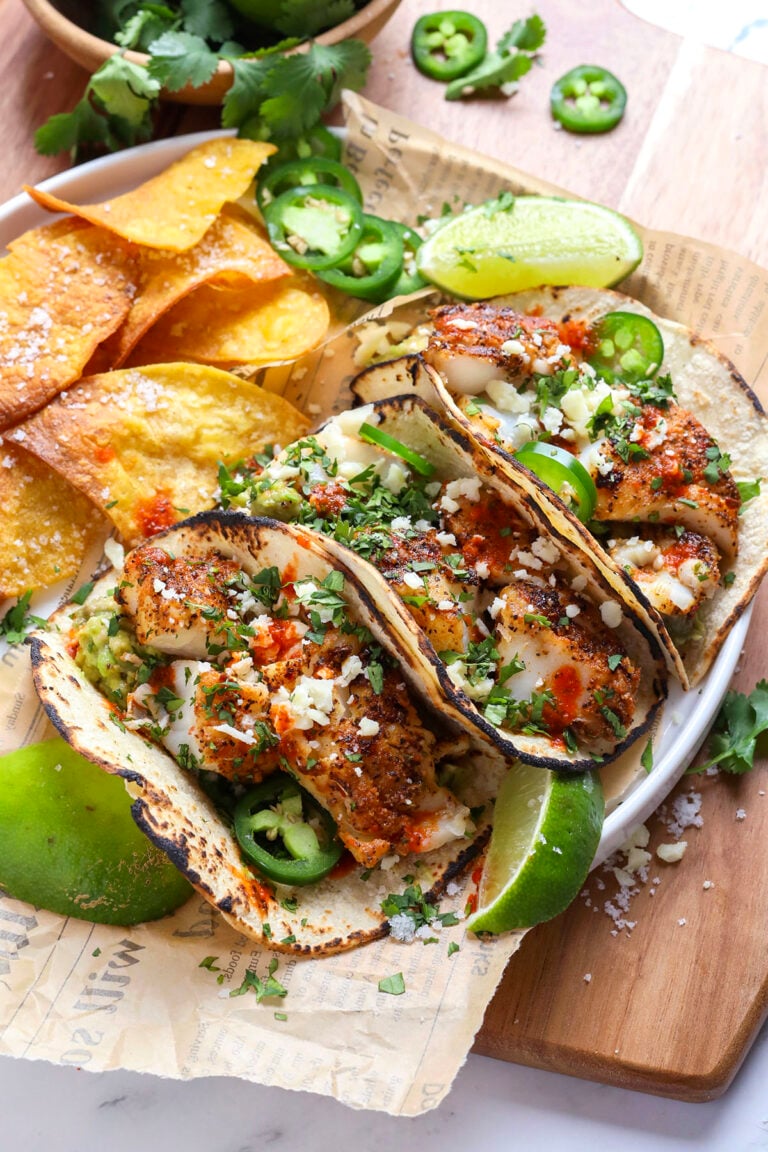 Three grilled fish tacos topped with chopped cilantro, jalapeños, cheese, and sauce, served with tortilla chips, lime wedges, and sliced jalapeños on a white plate with parchment paper, on a wooden board.