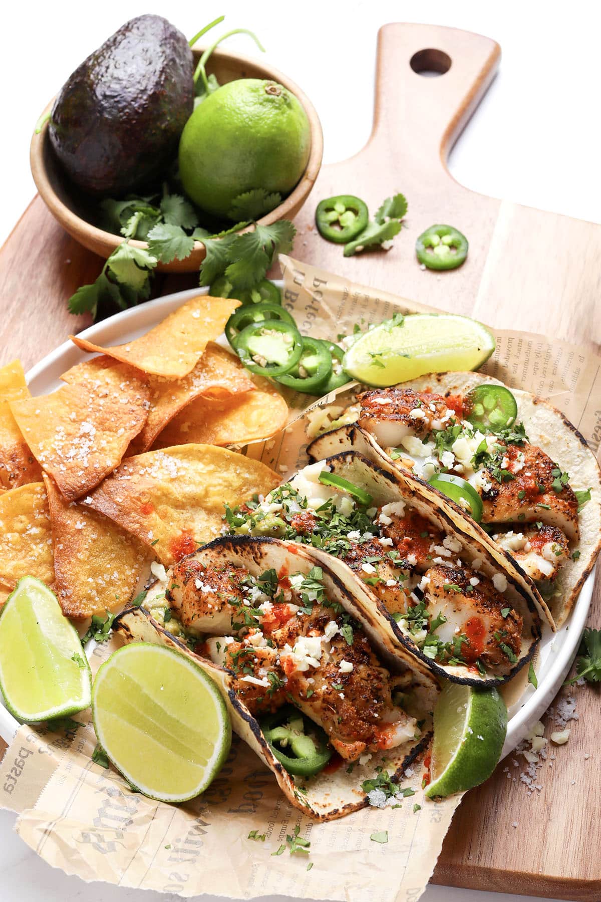 A plate of grilled fish tacos topped with diced vegetables, fresh cilantro, jalapeño slices, and cheese, served with lime wedges and tortilla chips on a wooden board beside an avocado, a lime, and cilantro.