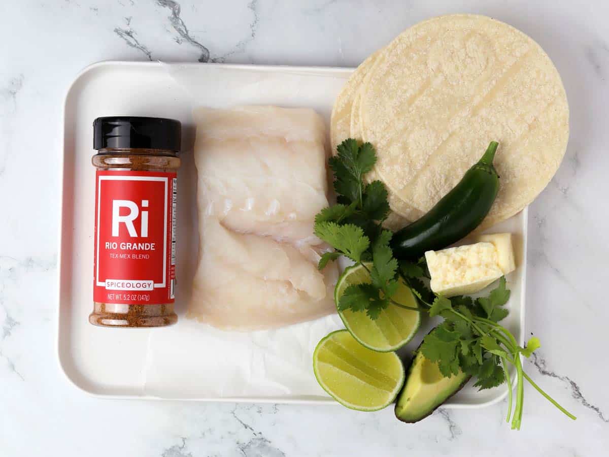 A tray with a jar of Rio Grande Tex-Mex spice blend, two raw fish fillets, corn tortillas, a jalapeño, cilantro, lime wedges, avocado slices, and a wedge of cheese on a marble surface.