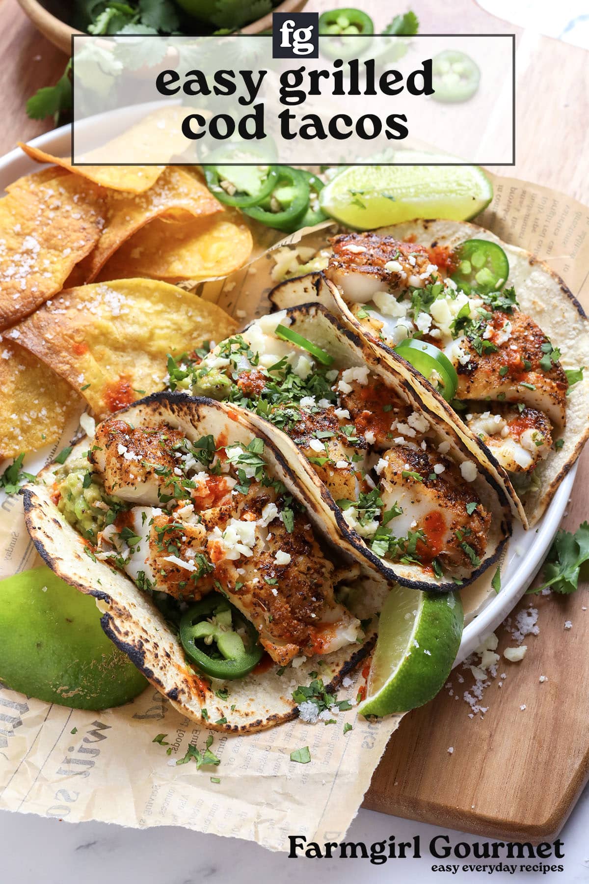 A plate of grilled cod tacos topped with salsa, cheese, cilantro, and jalapeño slices, served with lime wedges and crispy tortilla chips on the side. The text reads easy grilled cod tacos and Farmgirl Gourmet.