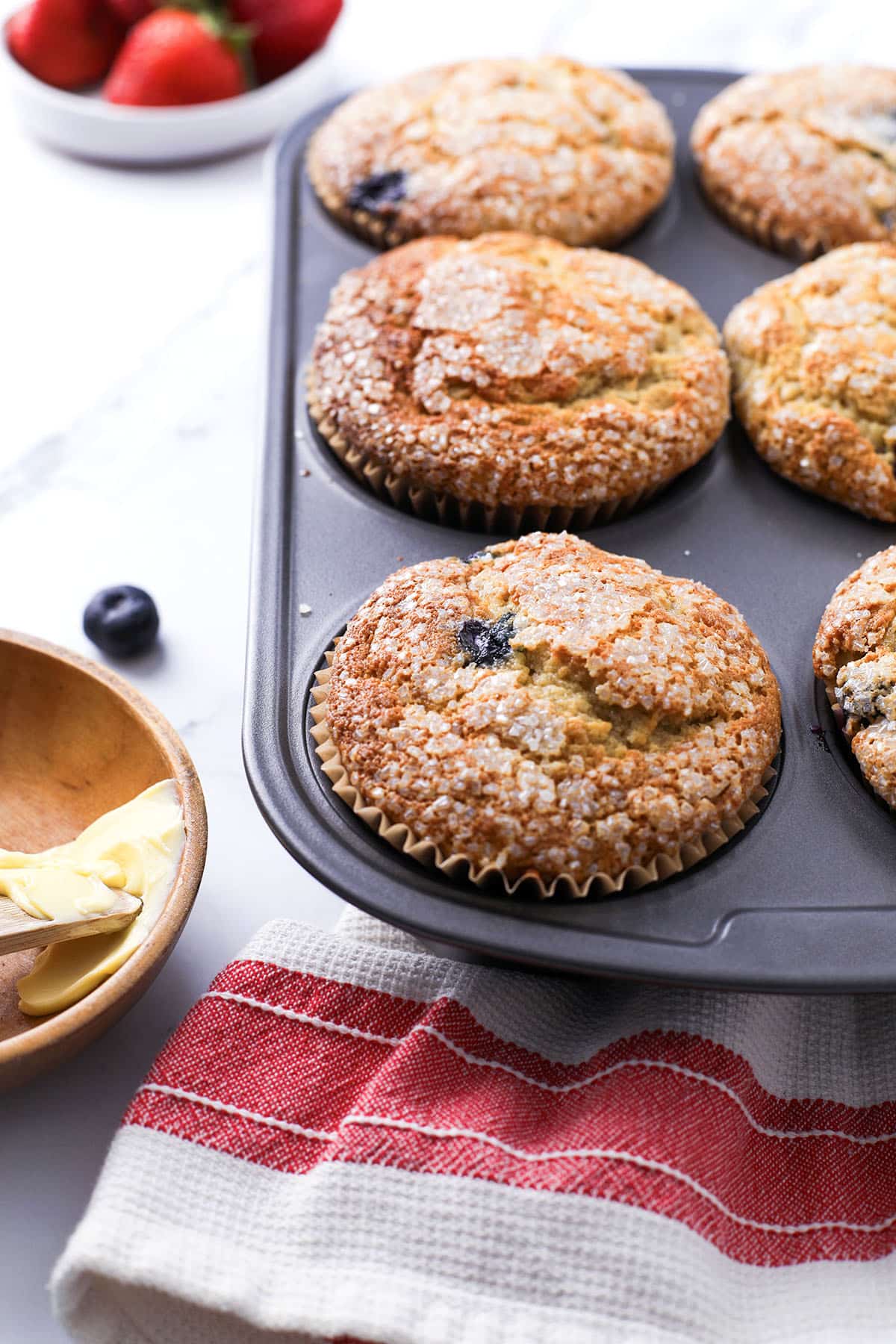 A muffin tin filled with golden-brown blueberry muffins topped with sugar crystals, next to a wooden bowl with butter, a red-striped towel, and a bowl of fresh strawberries in the background.