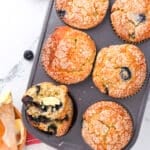 A muffin tin with six golden blueberry muffins, one split open with butter melting inside. Fresh strawberries and a wooden butter dish are nearby on a marble surface with a red-striped towel.