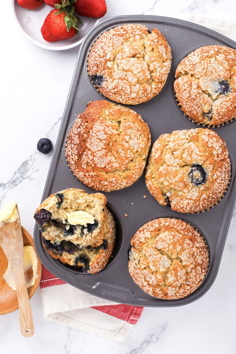 A muffin tin with six golden blueberry muffins, one split open with butter melting inside. Fresh strawberries and a wooden butter dish are nearby on a marble surface with a red-striped towel.