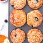 A muffin tin with six golden-brown muffins, some containing visible blueberries, sprinkled with sugar on top. Nearby are strawberries, a bowl of butter with a wooden knife, and a red-and-white towel on a marble countertop.