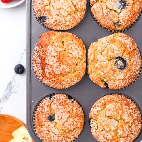 A muffin tin with six golden-brown muffins, some containing visible blueberries, sprinkled with sugar on top. Nearby are strawberries, a bowl of butter with a wooden knife, and a red-and-white towel on a marble countertop.