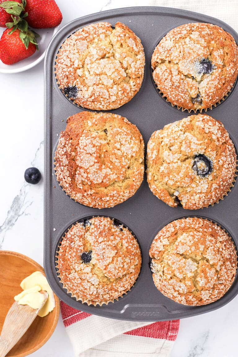 A muffin tin with six golden-brown muffins, some containing visible blueberries, sprinkled with sugar on top. Nearby are strawberries, a bowl of butter with a wooden knife, and a red-and-white towel on a marble countertop.