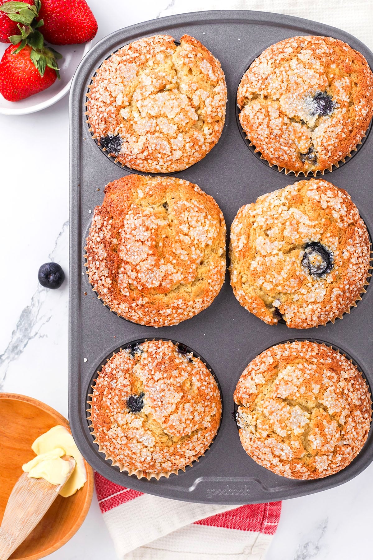 A muffin tin with six golden-brown muffins, some containing visible blueberries, sprinkled with sugar on top. Nearby are strawberries, a bowl of butter with a wooden knife, and a red-and-white towel on a marble countertop.