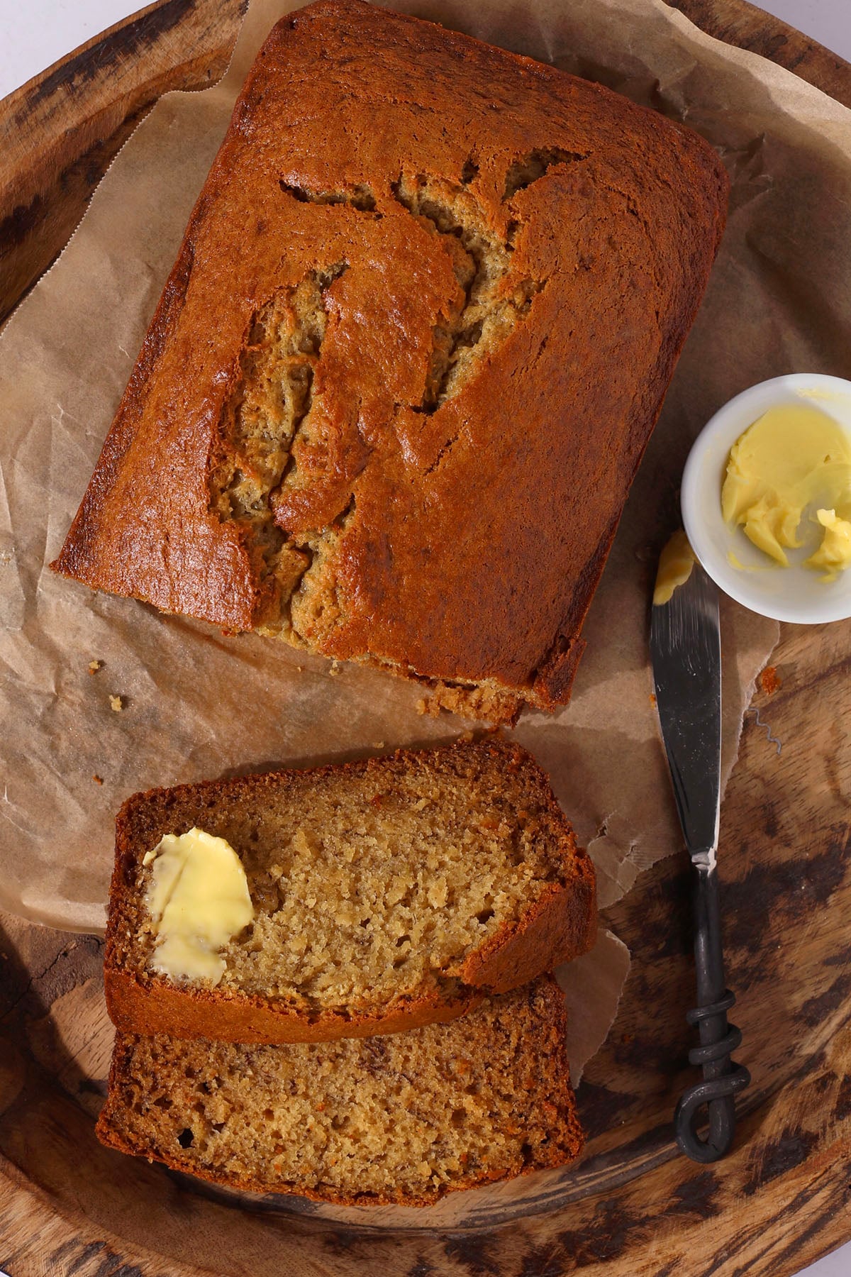 A loaf of banana bread on parchment paper with two slices cut, one spread with butter. A knife and a small bowl of butter are placed beside the bread on a wooden tray.
