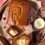 A loaf of banana bread on parchment paper, partially sliced, with two slices spread with butter. A butter knife and a small dish of butter are beside the bread, all on a wooden tray atop a pink tiled surface.