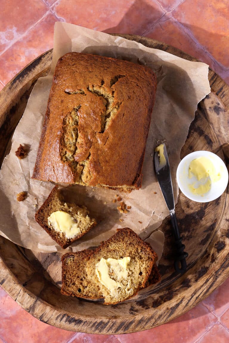 A loaf of banana bread on parchment paper, partially sliced, with two slices spread with butter. A butter knife and a small dish of butter are beside the bread, all on a wooden tray atop a pink tiled surface.