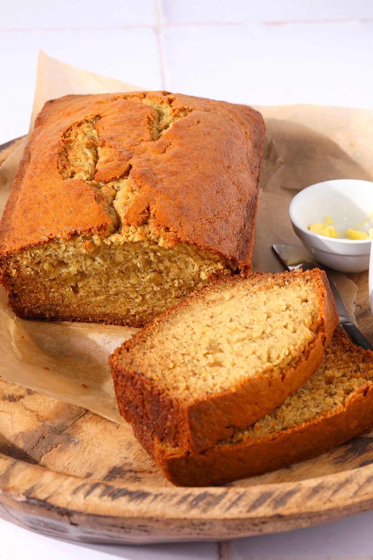 A loaf of banana bread sits on parchment paper atop a wooden tray, with two slices cut and laid in front. A small white bowl with butter and a knife are beside the bread.