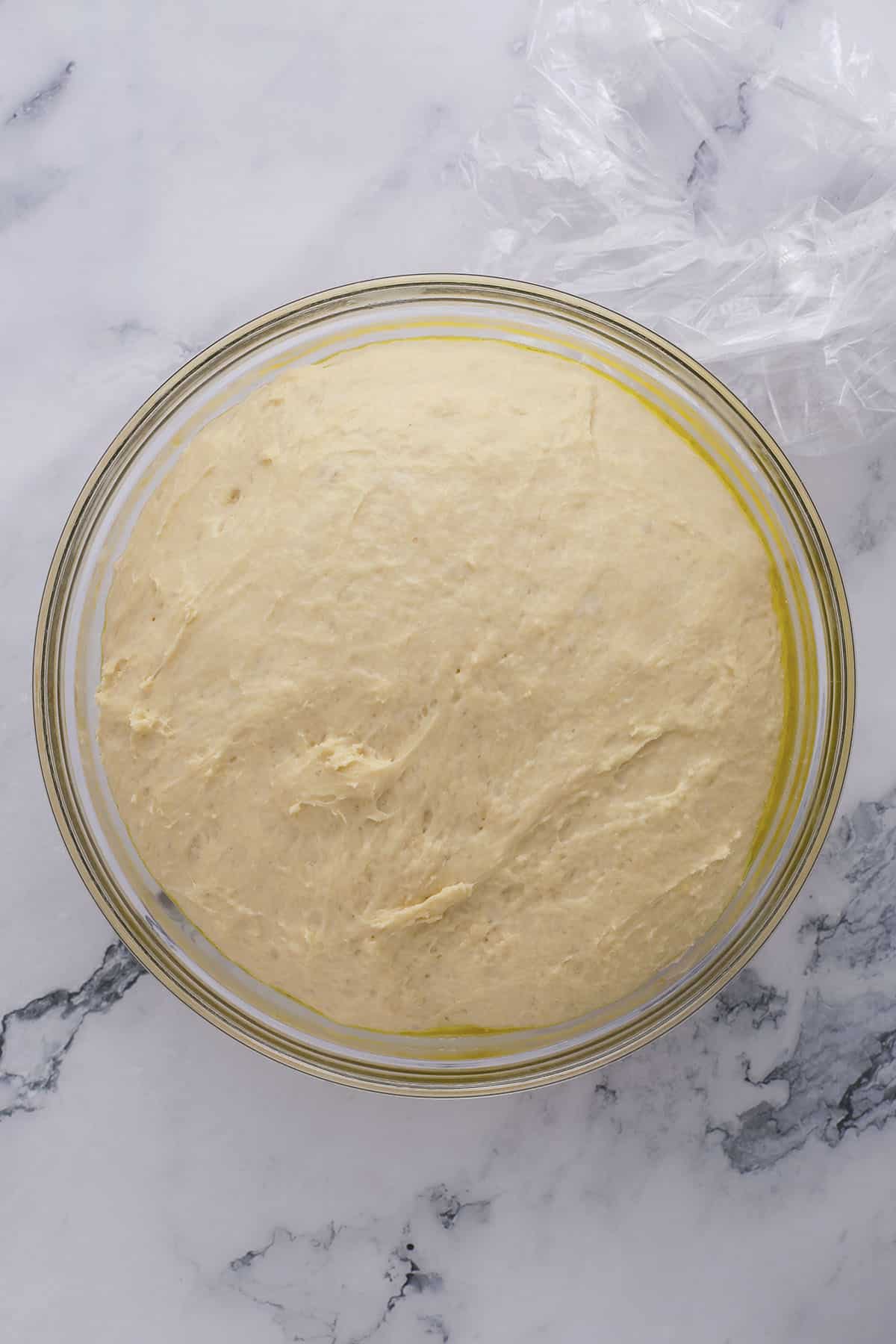 A glass bowl filled with risen bread dough sits on a white marble surface, with a piece of plastic wrap partially visible in the top right corner.