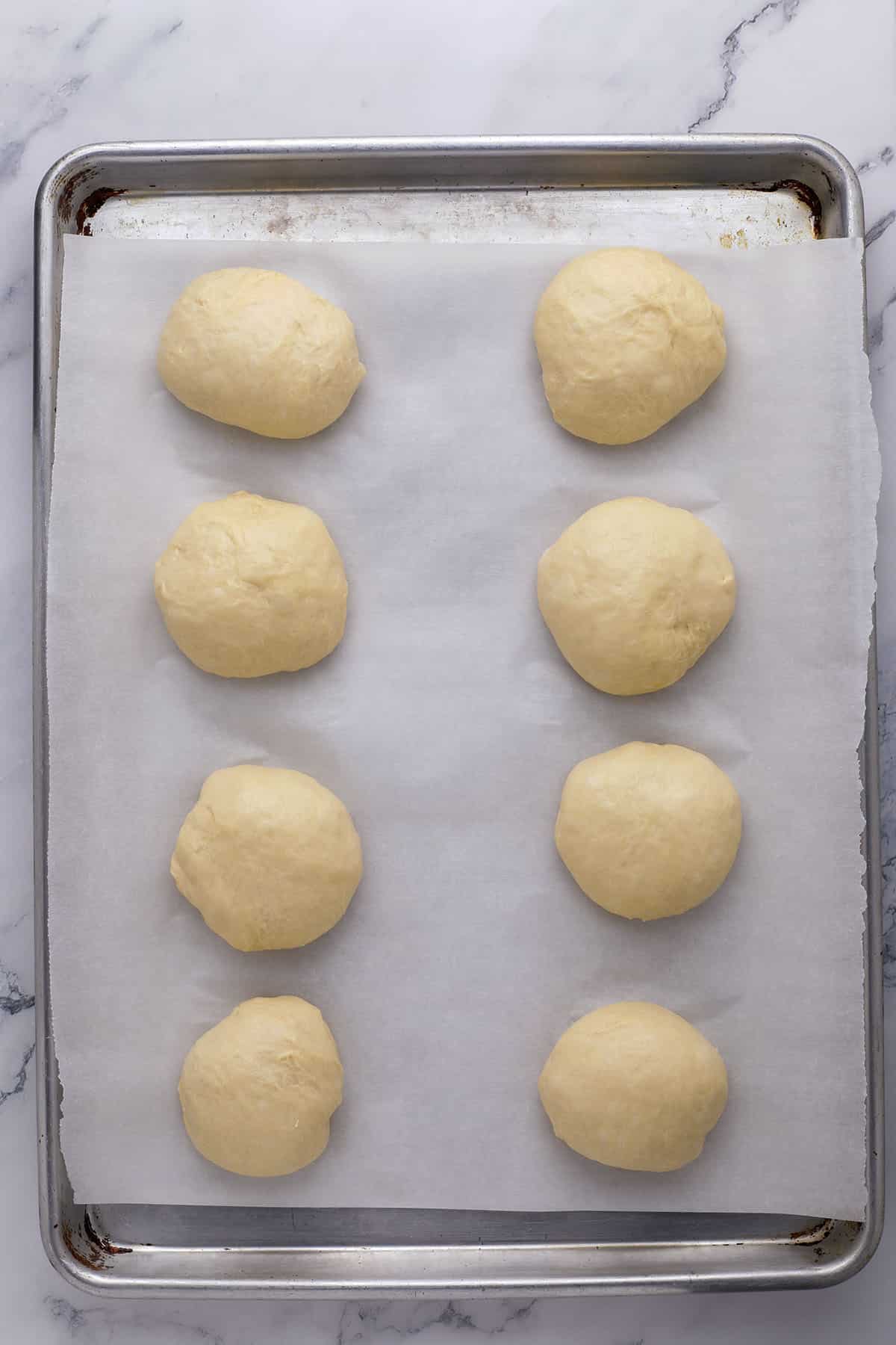 Eight round balls of dough are evenly spaced on a parchment-lined baking sheet, resting on a marble countertop, ready to be baked.