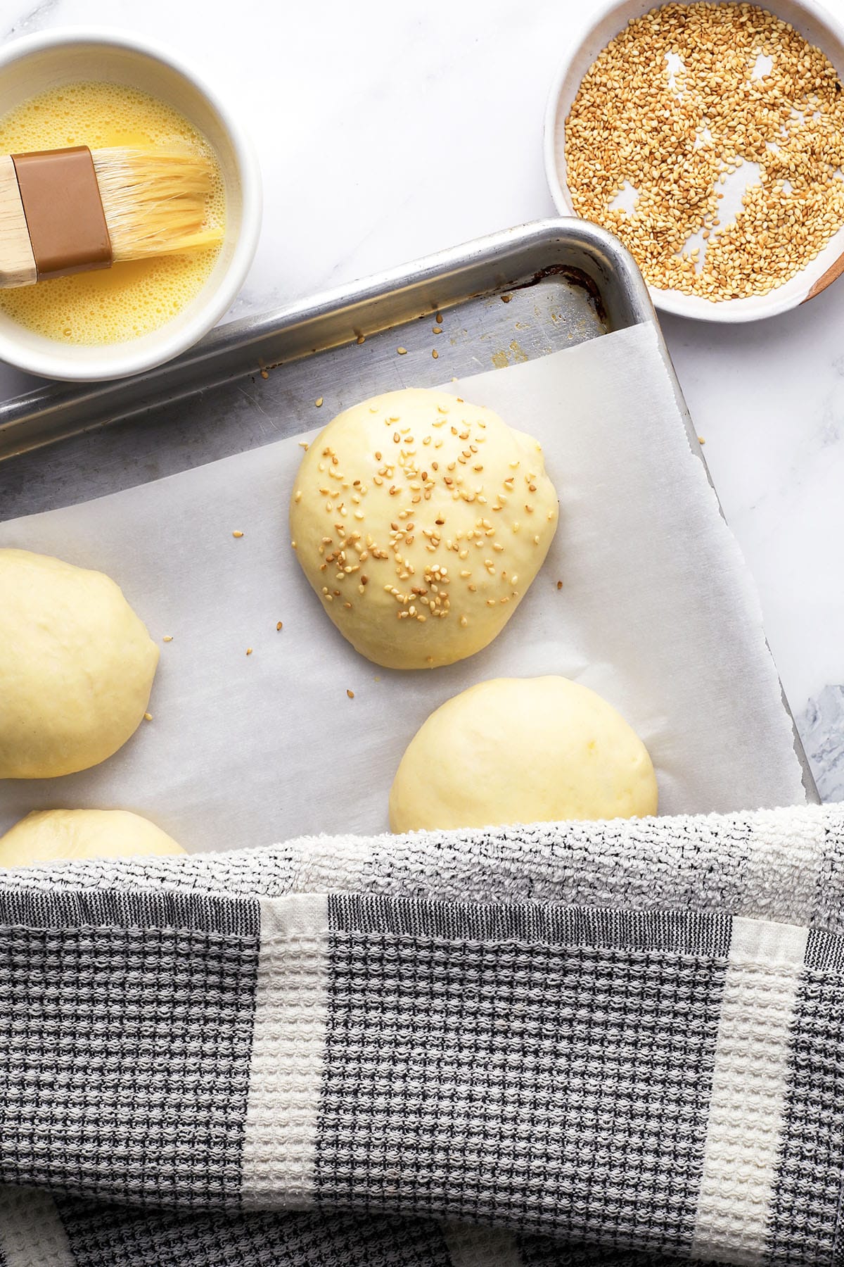 A baking tray with round dough balls on parchment paper, one topped with sesame seeds. A bowl of beaten egg with a brush and a bowl of sesame seeds are nearby. A checkered kitchen towel partly covers the tray.