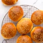 Seven golden brown sesame seed buns are cooling on a round wire rack, with one additional bun placed on a white marble surface beside the rack. The buns have a shiny finish and are evenly spaced.