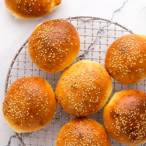 Seven golden brown sesame seed buns are cooling on a round wire rack, with one additional bun placed on a white marble surface beside the rack. The buns have a shiny finish and are evenly spaced.