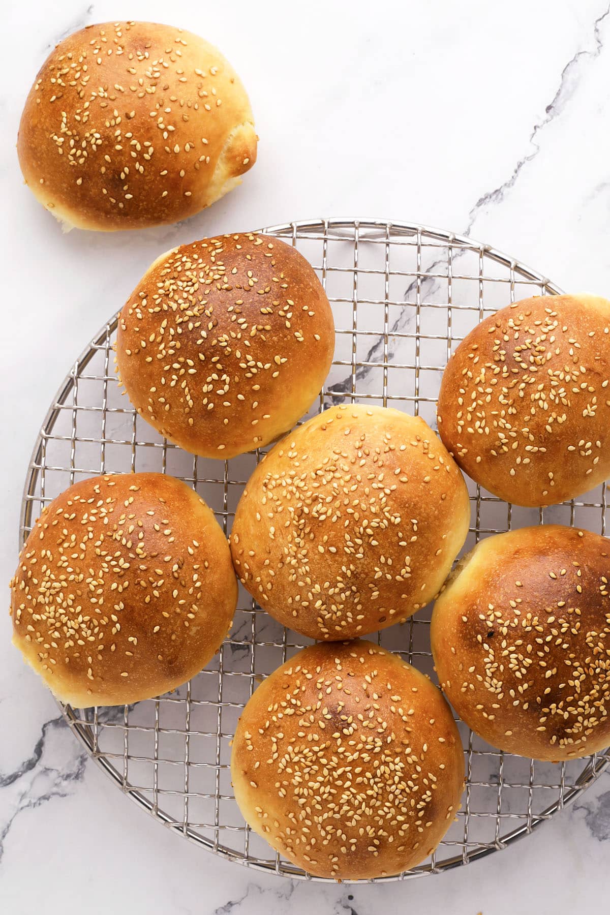 Seven golden brown sesame seed buns are cooling on a round wire rack, with one additional bun placed on a white marble surface beside the rack. The buns have a shiny finish and are evenly spaced.