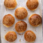 Eight golden brown sesame seed hamburger buns on a parchment paper-lined baking sheet, viewed from above. Some seeds are scattered around the buns on the paper.