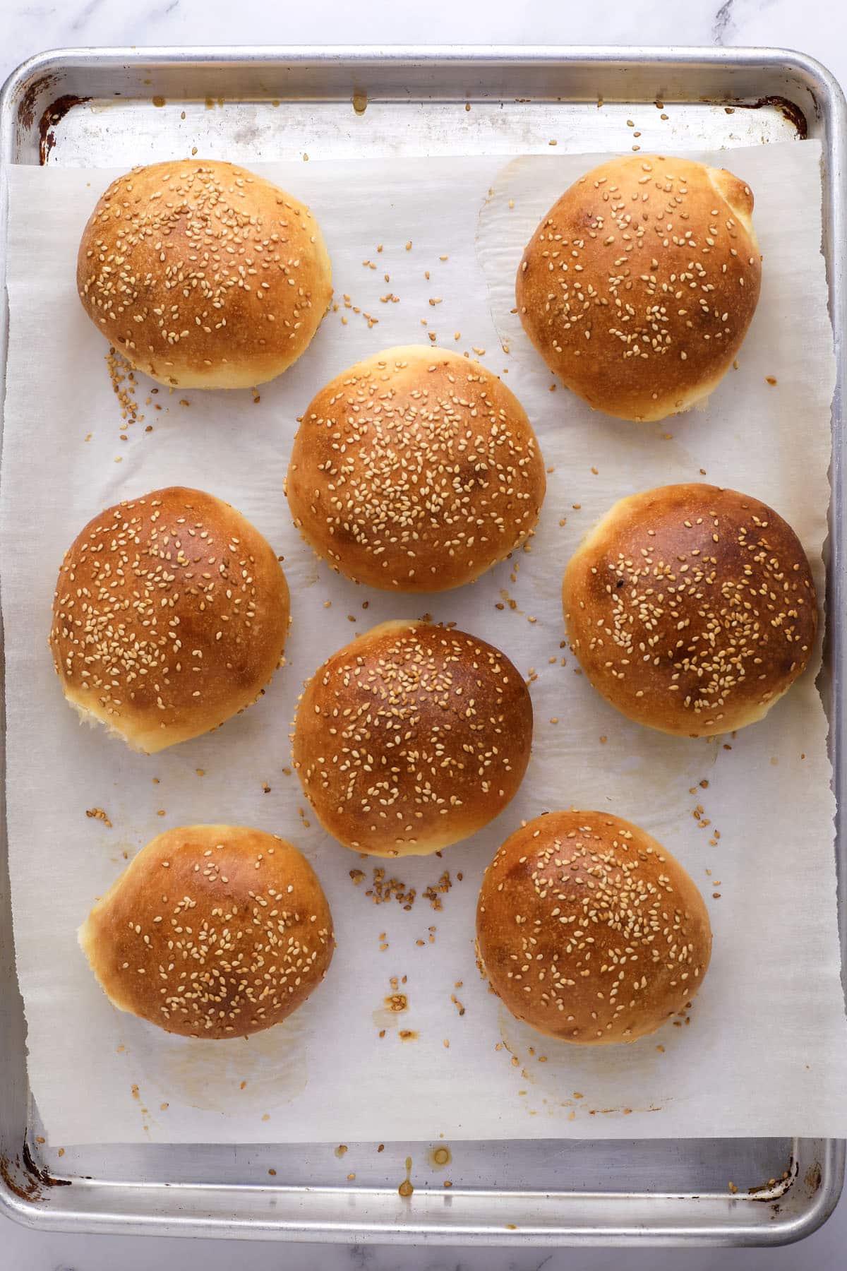 Eight golden brown sesame seed hamburger buns on a parchment paper-lined baking sheet, viewed from above. Some seeds are scattered around the buns on the paper.