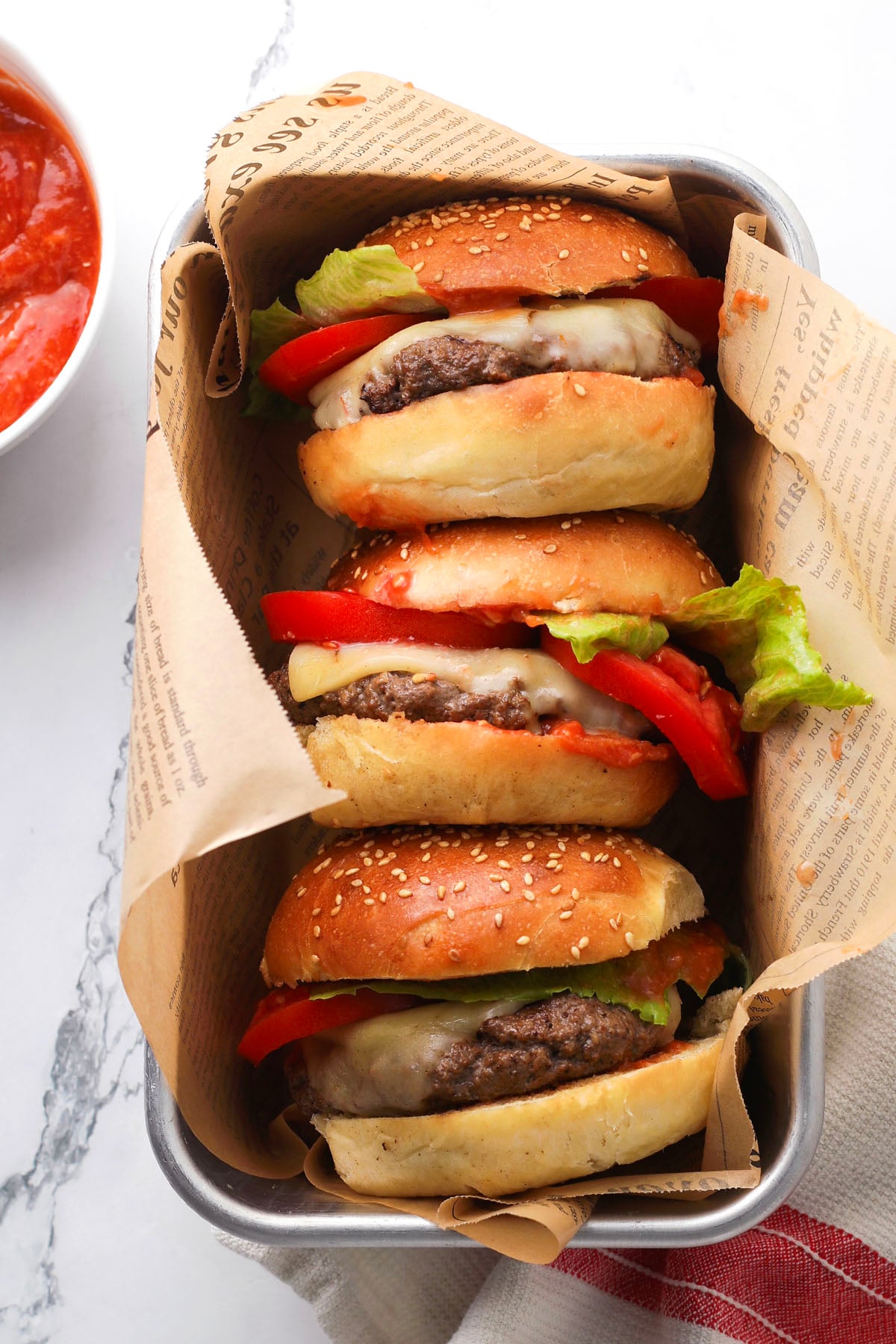 Three cheeseburgers with lettuce and tomato are stacked in a metal tray lined with newspaper-style paper; a bowl of red sauce is partially visible on the side.