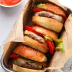 Three cheeseburgers with lettuce, tomato, and melted cheese in sesame buns are wrapped in newspaper-style paper and placed in a tray. A bowl of red sauce with a spoon is nearby on a marble surface.