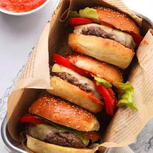 Three cheeseburgers with lettuce, tomato, and melted cheese in sesame buns are wrapped in newspaper-style paper and placed in a tray. A bowl of red sauce with a spoon is nearby on a marble surface.