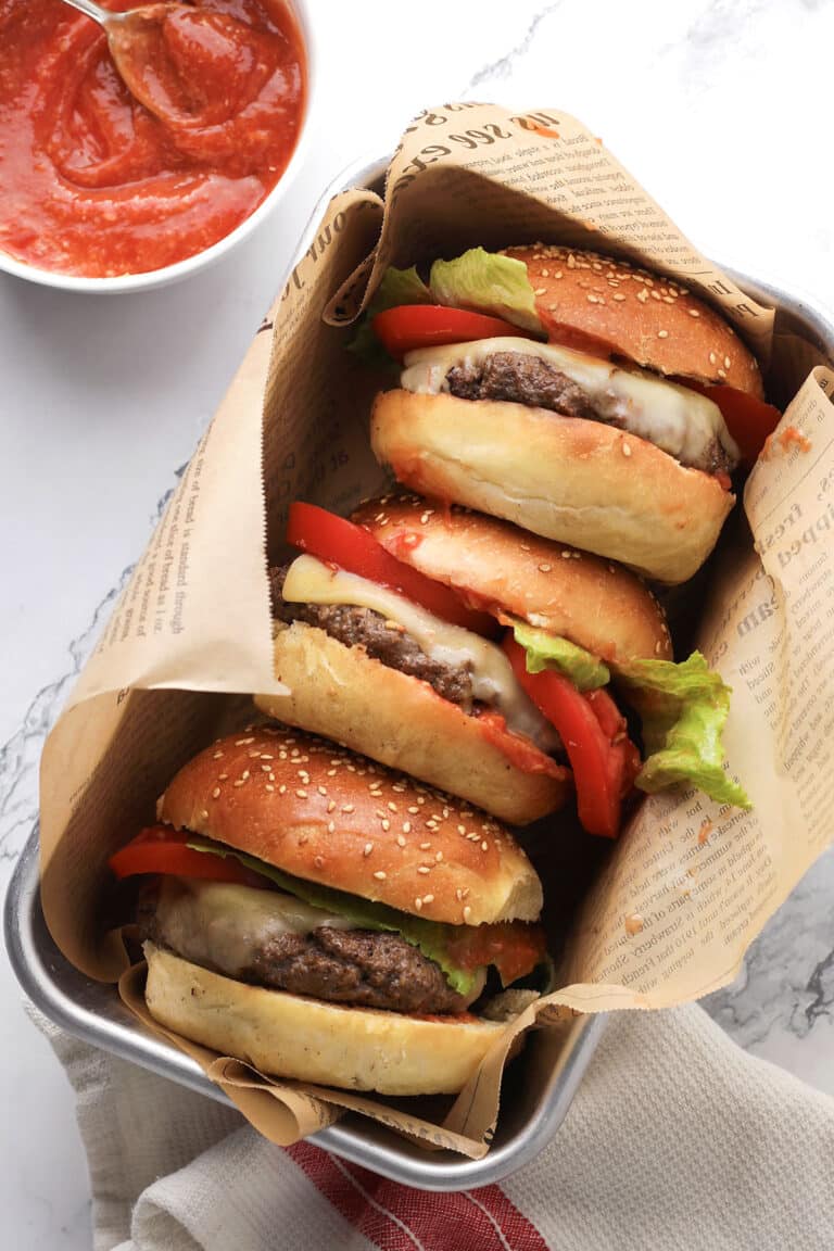 Three cheeseburgers with lettuce, tomato, and melted cheese in sesame buns are wrapped in newspaper-style paper and placed in a tray. A bowl of red sauce with a spoon is nearby on a marble surface.