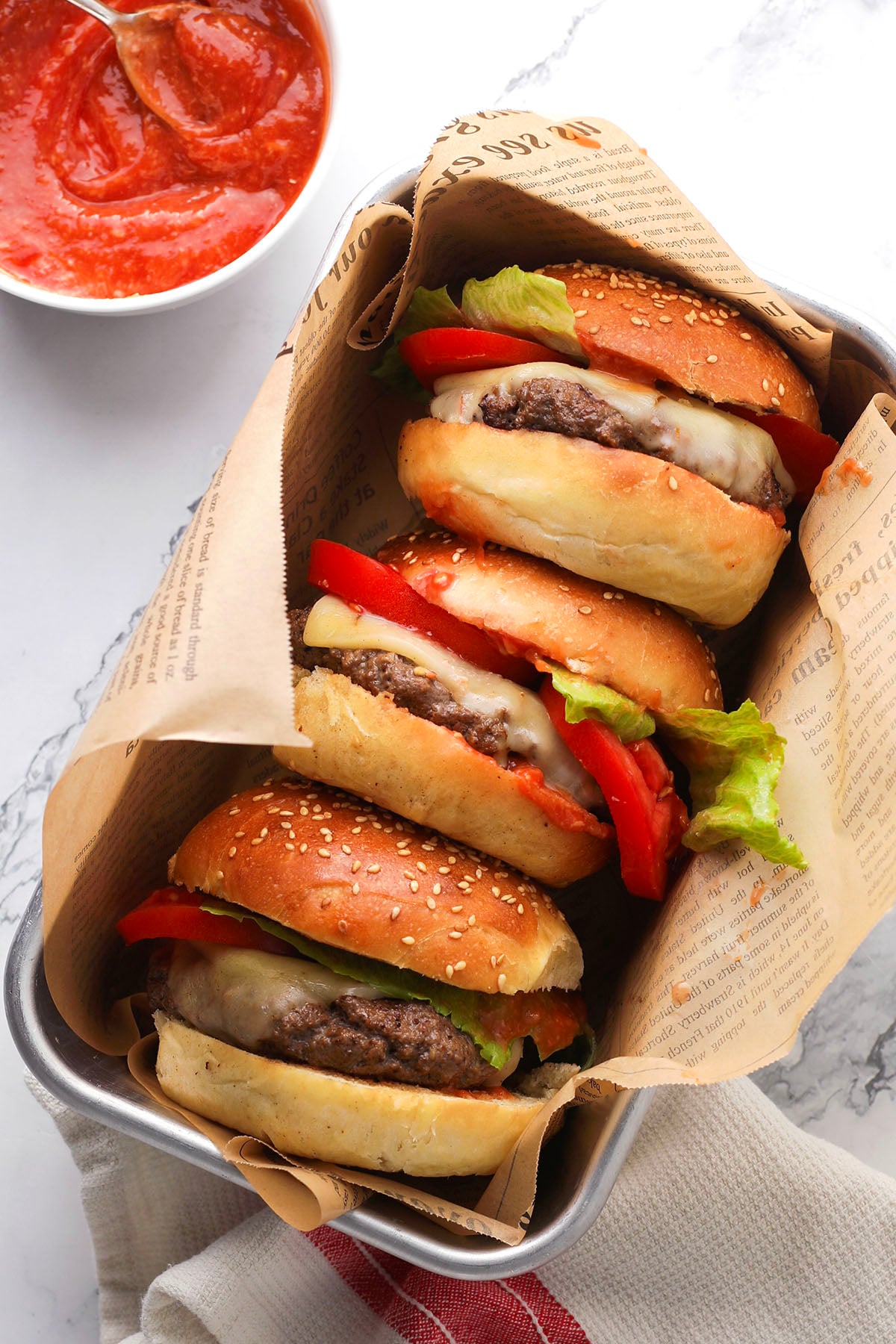 Three cheeseburgers with lettuce, tomato, and melted cheese in sesame buns are wrapped in newspaper-style paper and placed in a tray. A bowl of red sauce with a spoon is nearby on a marble surface.