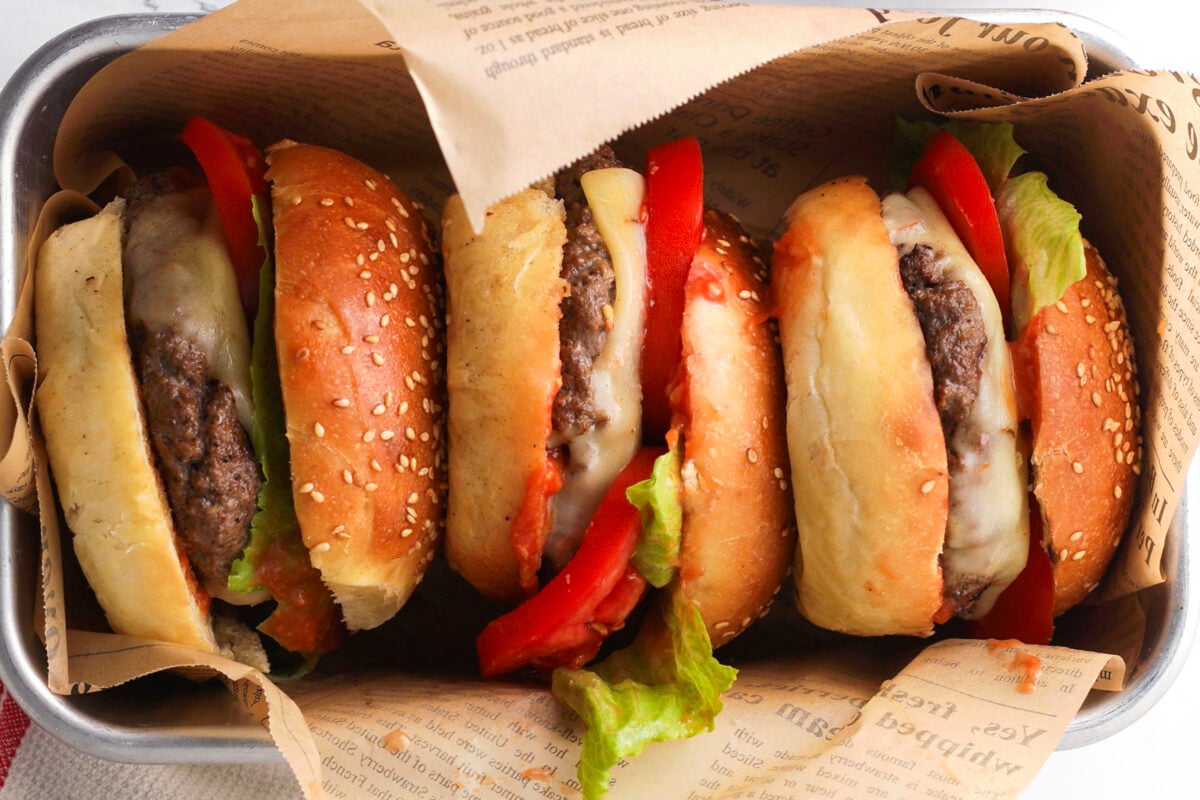 Three cheeseburgers with lettuce, tomato, melted cheese, and beef patties in sesame seed buns are arranged in a metal tray lined with newspaper-style parchment paper.