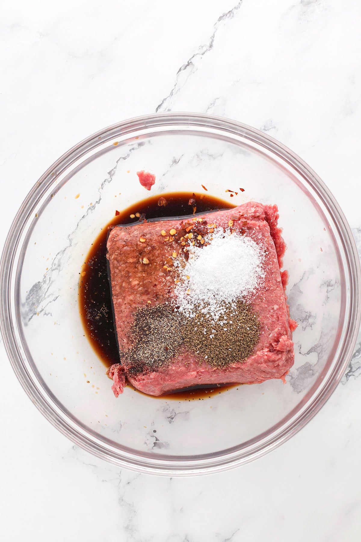 A glass bowl on a marble surface filled with raw ground beef, soy sauce, red pepper flakes, salt, and black pepper, ready to be mixed.
