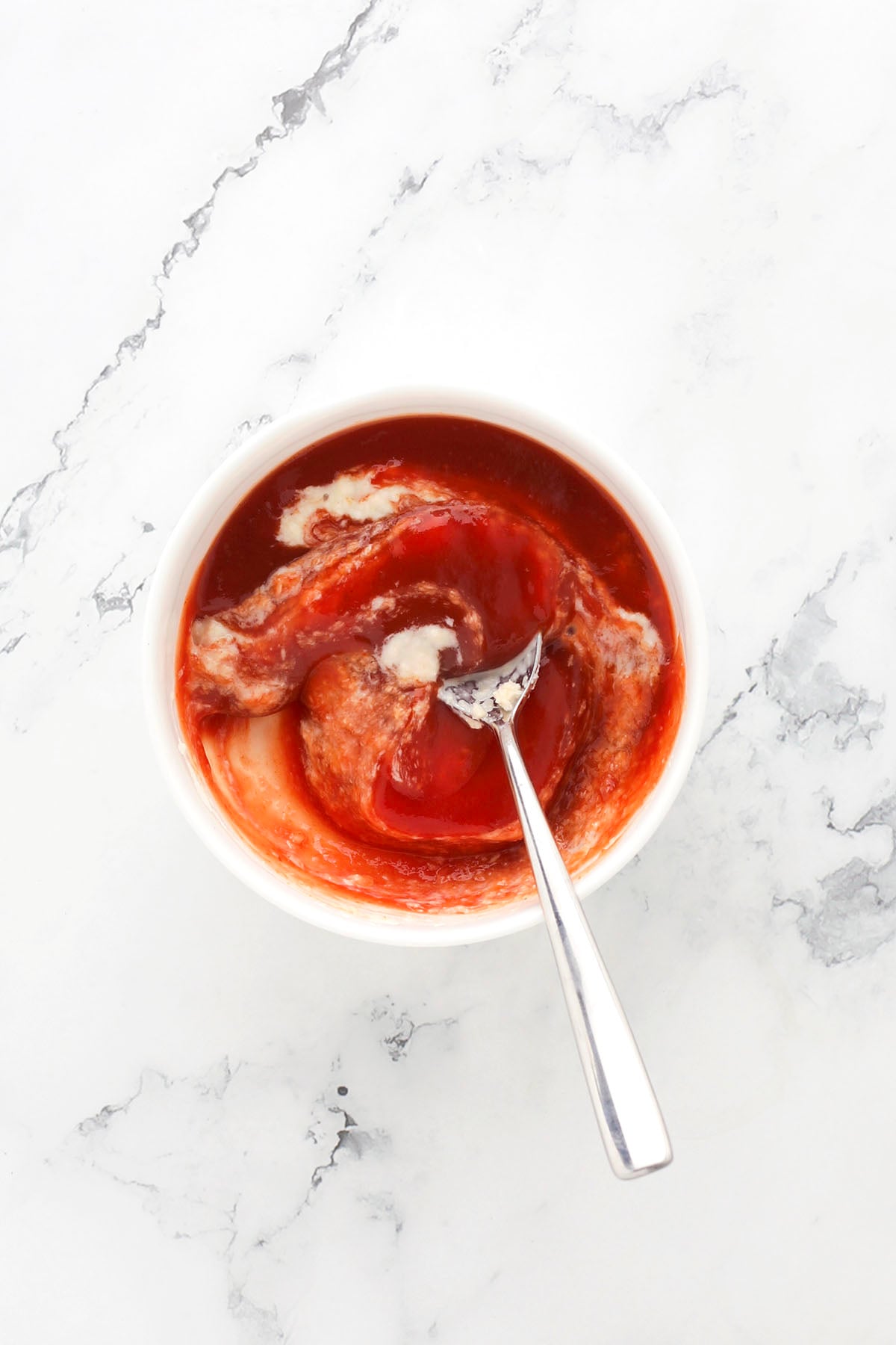A white bowl filled with ketchup, mayonnaise, and mustard being mixed together with a spoon, set on a white marble surface.